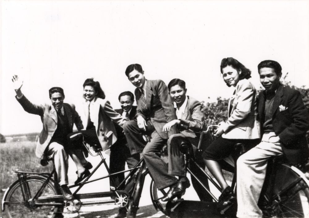 Tujuh anak muda Indonesia berjejer di atas sepeda tandem panjang di sebuah padang di Belanda, tertawa ke arah kamera, foto hitam putih tahun 1942.